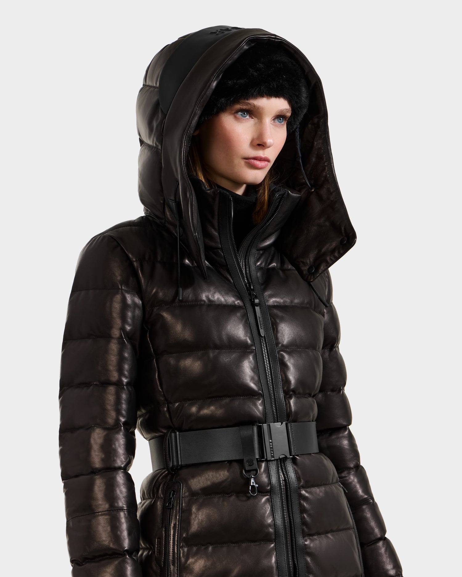 A woman wears the Rudsak PELIN X - 8125575 Black leather down puffer jacket with hood and belted waist, paired with a black fur hat, against a plain white background.