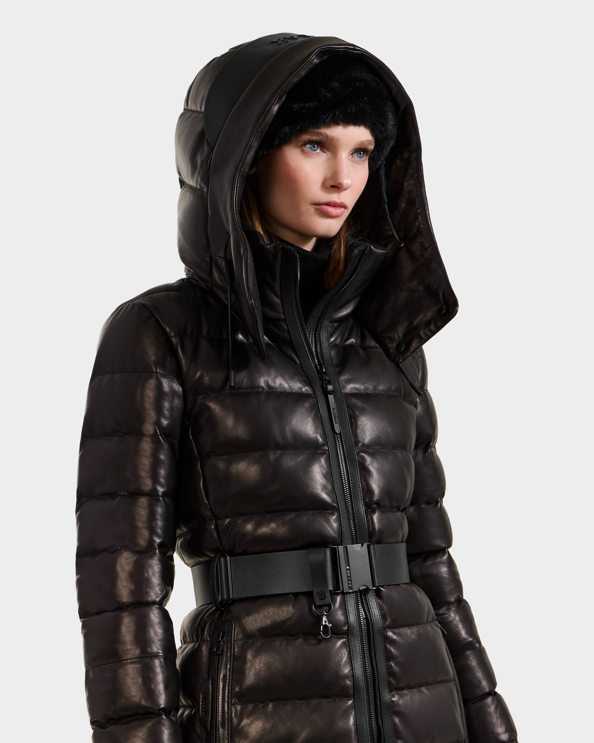 A woman wears the Rudsak PELIN X - 8125575 Black leather down puffer jacket with hood and belted waist, paired with a black fur hat, against a plain white background.
