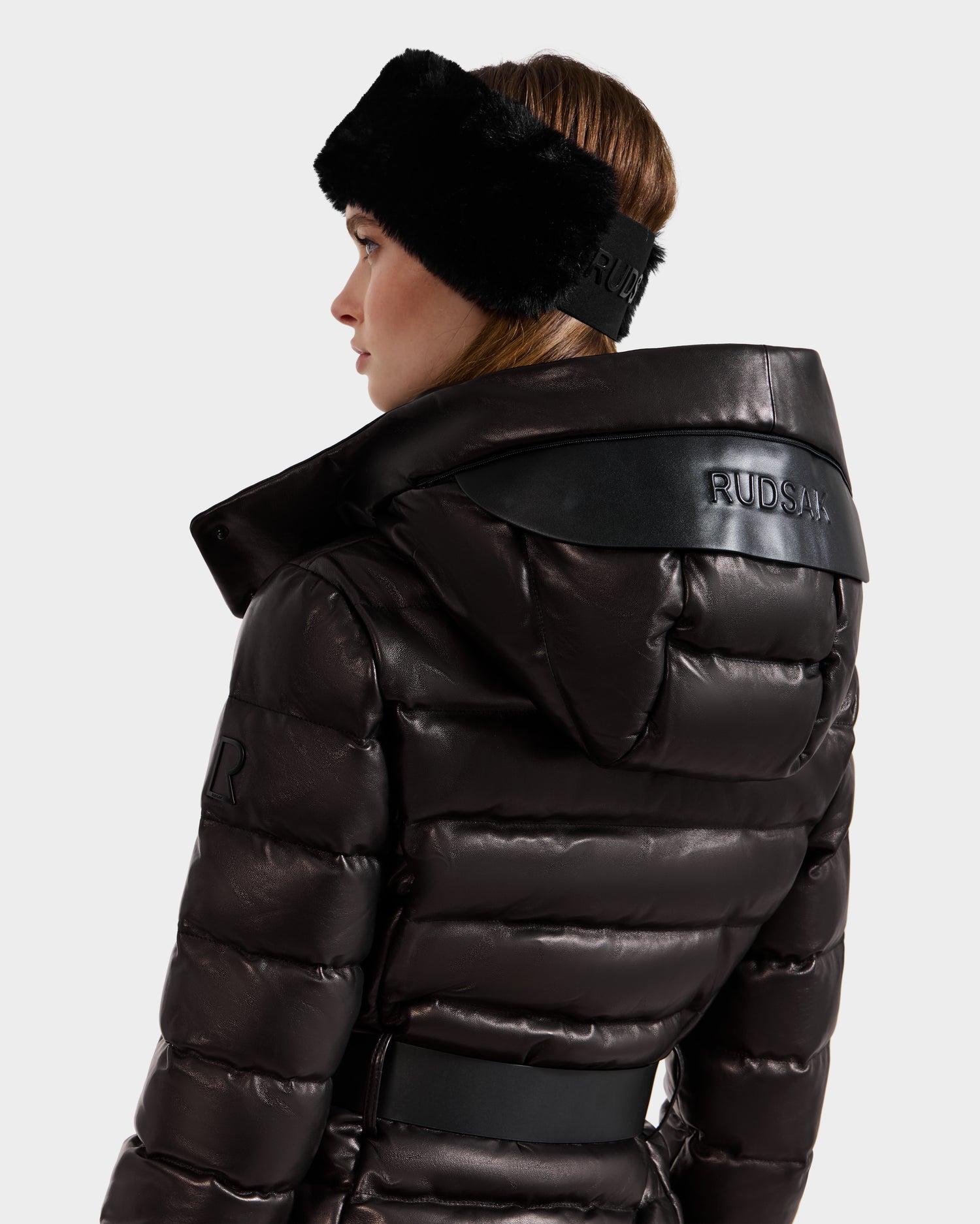 A woman wearing the Rudsak PELIN X - 8125575 Black leather down puffer with the brand embossed on the back and a matching black faux fur headband, shown from behind and in profile against a white background.