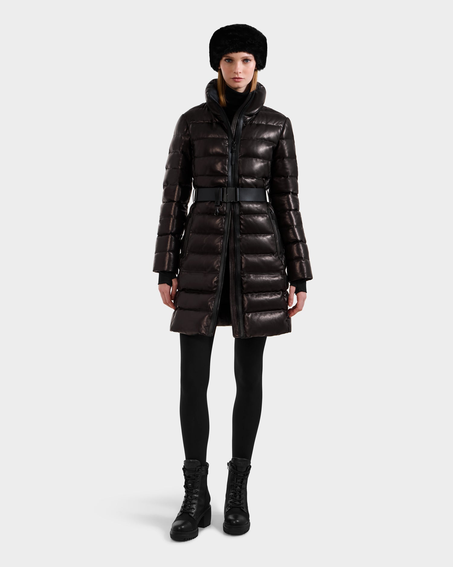 A woman stands facing forward in the Rudsak PELIN X - 8125575 Black leather down puffer with a belt, paired with black leggings, lace-up boots, and a black fur hat against a plain light background.