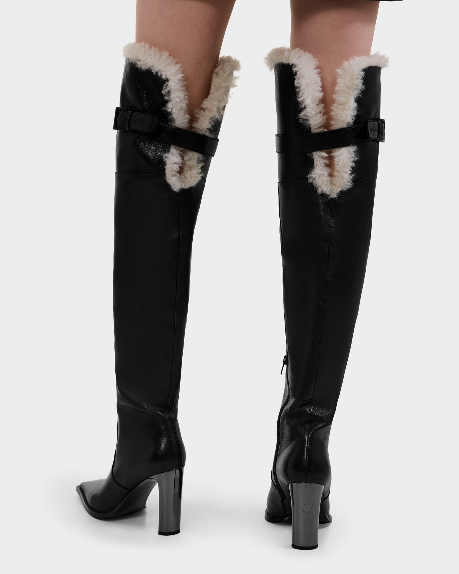 A person wears Rudsak PERNA - 8225555 Black knee-high heeled leather boots with white fur trim and back buckles, shown from behind against a plain background—the perfect women's winter boots for a stylish look.