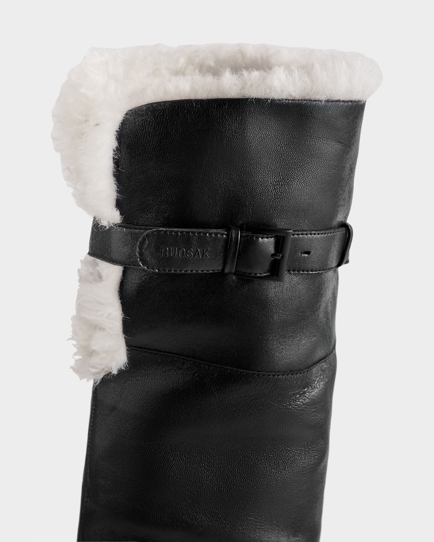 Close-up of the PERNA - 8225555 Black tall leather boot by Rudsak, featuring a white shearling cuff and a decorative black strap with buckle around the upper part.