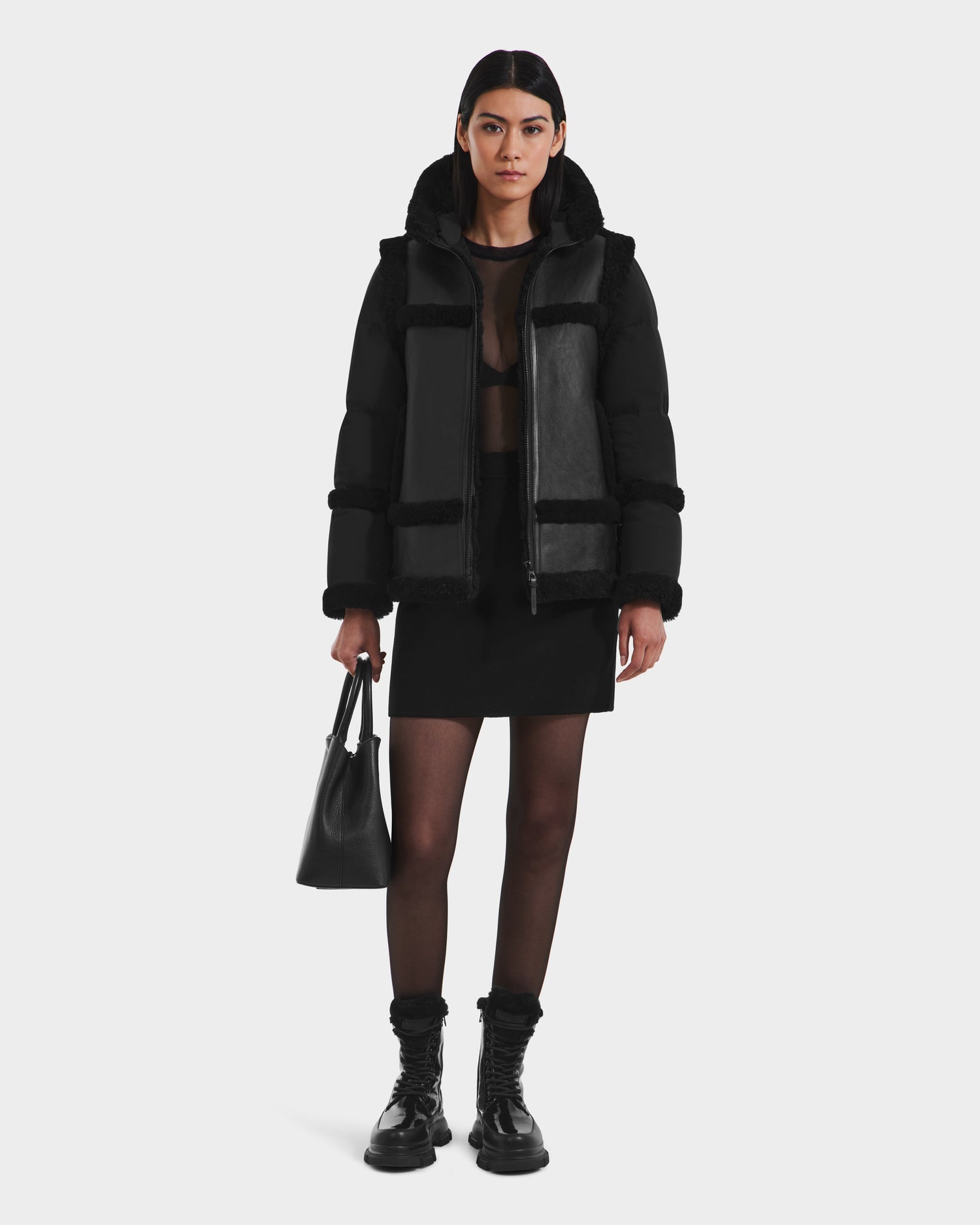 A woman with straight dark hair and a neutral expression wears the Rudsak PHEOBE - 8125510 Black shearling jacket as transitional outerwear over a sheer top, skirt, tights, and combat boots, holding a black handbag.