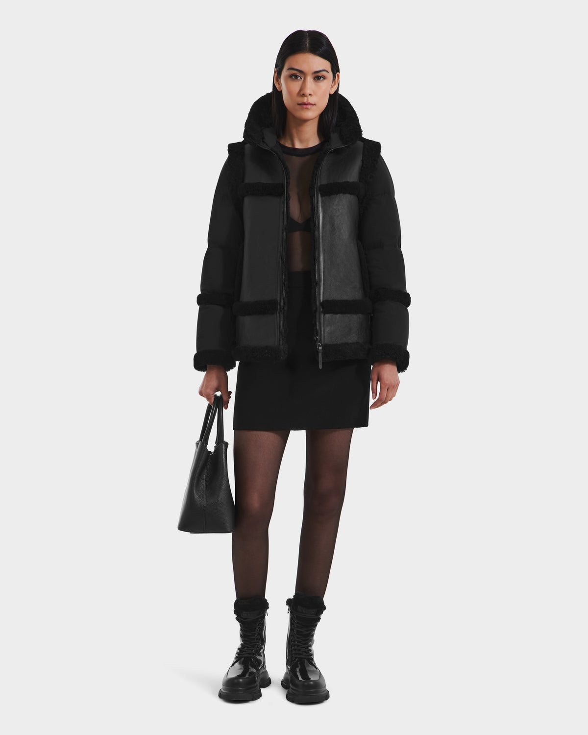 A woman with straight dark hair and a neutral expression wears the Rudsak PHEOBE - 8125510 Black shearling jacket as transitional outerwear over a sheer top, skirt, tights, and combat boots, holding a black handbag.