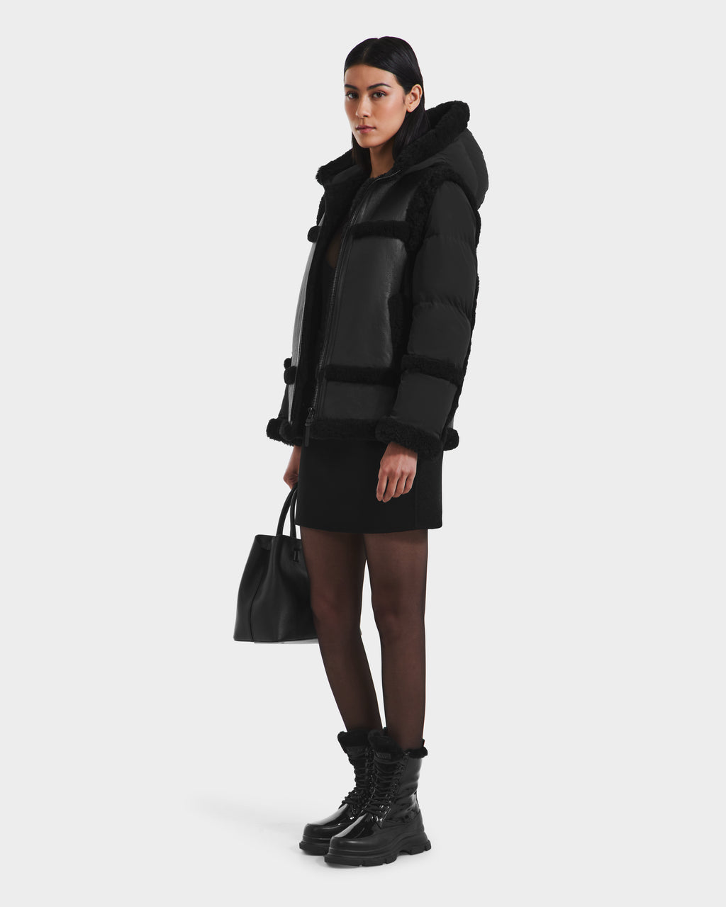 A woman wears the Rudsak PHEOBE - 8125510 Black puffer combo jacket with a fur-lined hood, paired with a black dress, sheer tights, lace-up boots, and holds a black handbag against a white background—a perfect transitional outerwear style.