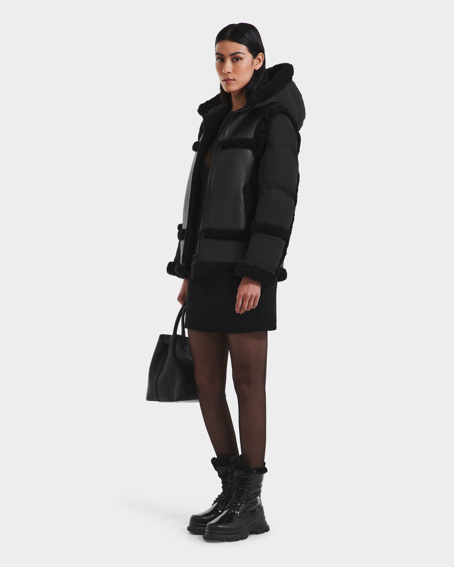 A woman wears the Rudsak PHEOBE - 8125510 Black puffer combo jacket with a fur-lined hood, paired with a black dress, sheer tights, lace-up boots, and holds a black handbag against a white background—a perfect transitional outerwear style.