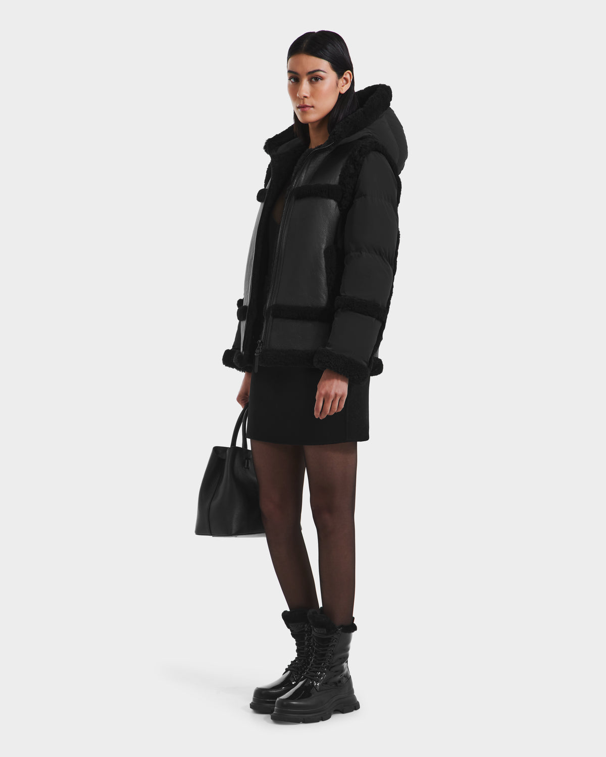 A woman wears the Rudsak PHEOBE - 8125510 Black puffer combo jacket with a fur-lined hood, paired with a black dress, sheer tights, lace-up boots, and holds a black handbag against a white background—a perfect transitional outerwear style.