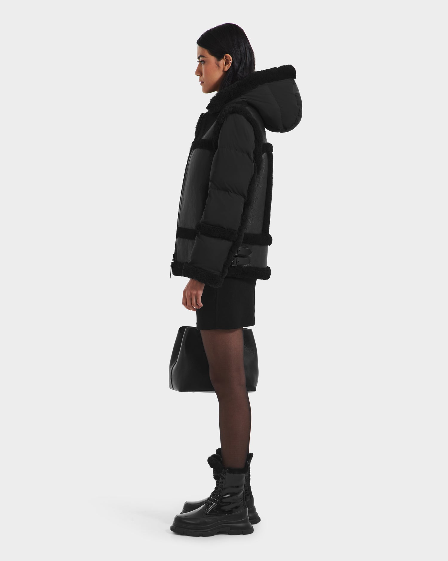 A woman stands in profile wearing the Rudsak PHEOBE - 8125510 Black down and feather insulated jacket with fur trim, styled with a skirt, sheer tights, boots, and a handbag against a light background.