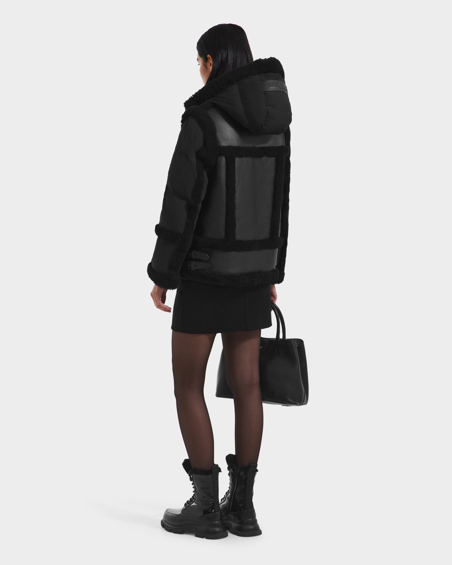 A person with long dark hair, wearing the Rudsak PHEOBE - 8125510 Black womens shearling puffer jacket, a black skirt, tights, and combat boots, stands facing away while holding a black handbag against a white background.