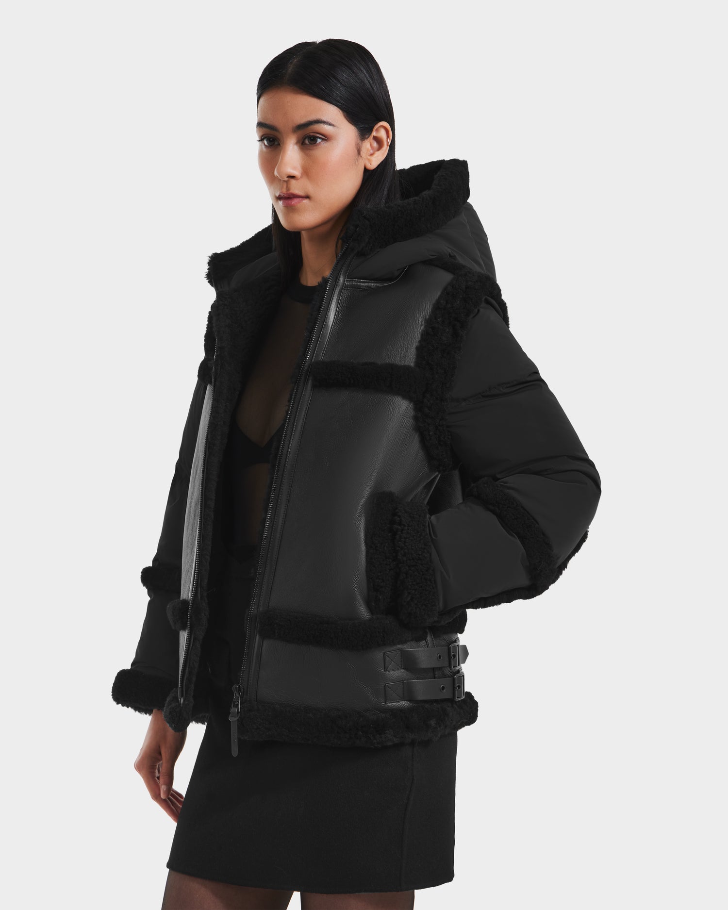A woman stands against a plain white background wearing the Rudsak PHEOBE - 8125510 Black women’s shearling jacket with faux fur trim, buckle details, and a short black skirt.
