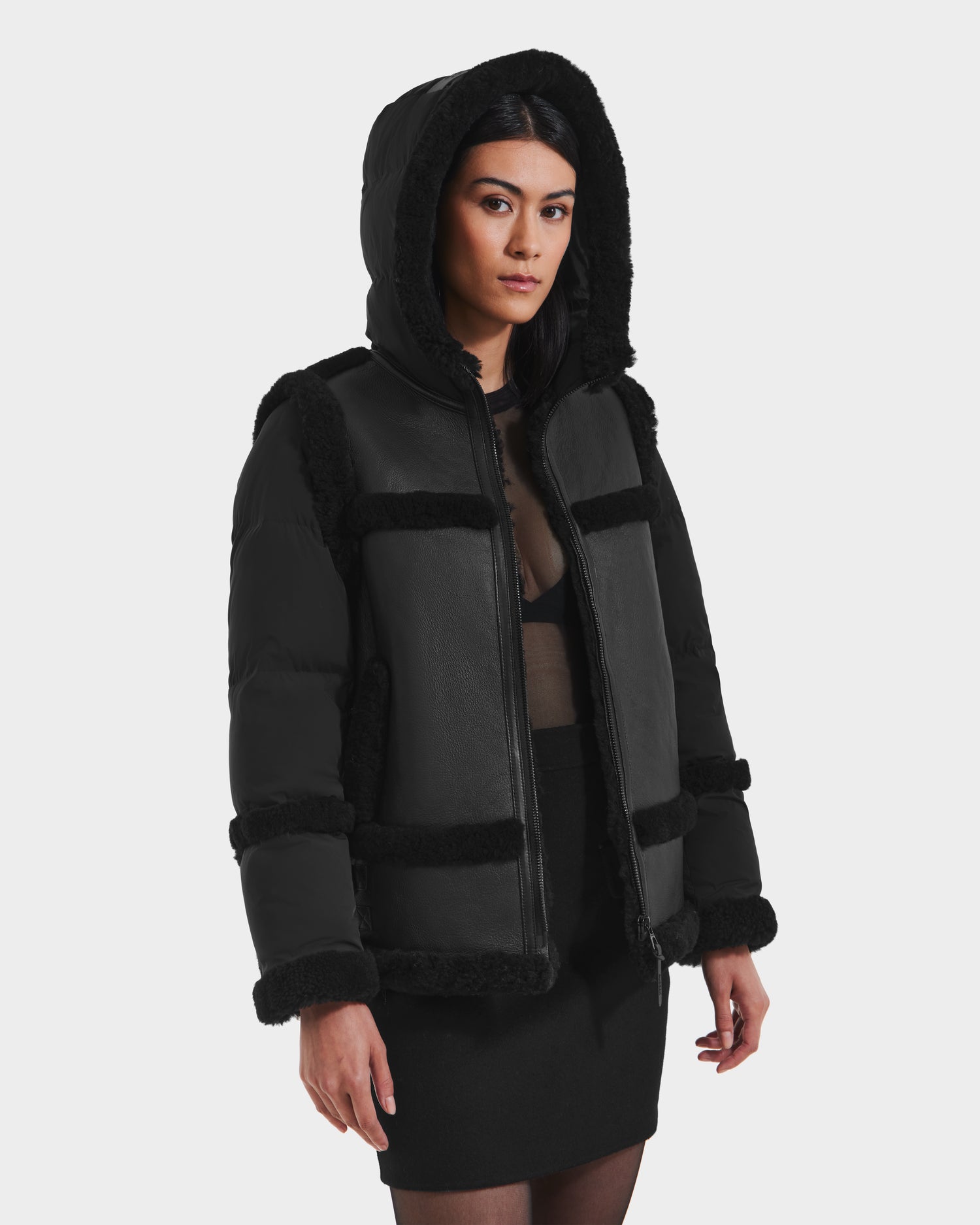 A woman wears the Rudsak PHEOBE - 8125510 Black women’s shearling puffer jacket over a sheer black top and skirt, standing against a white background and looking to the side with a neutral expression.