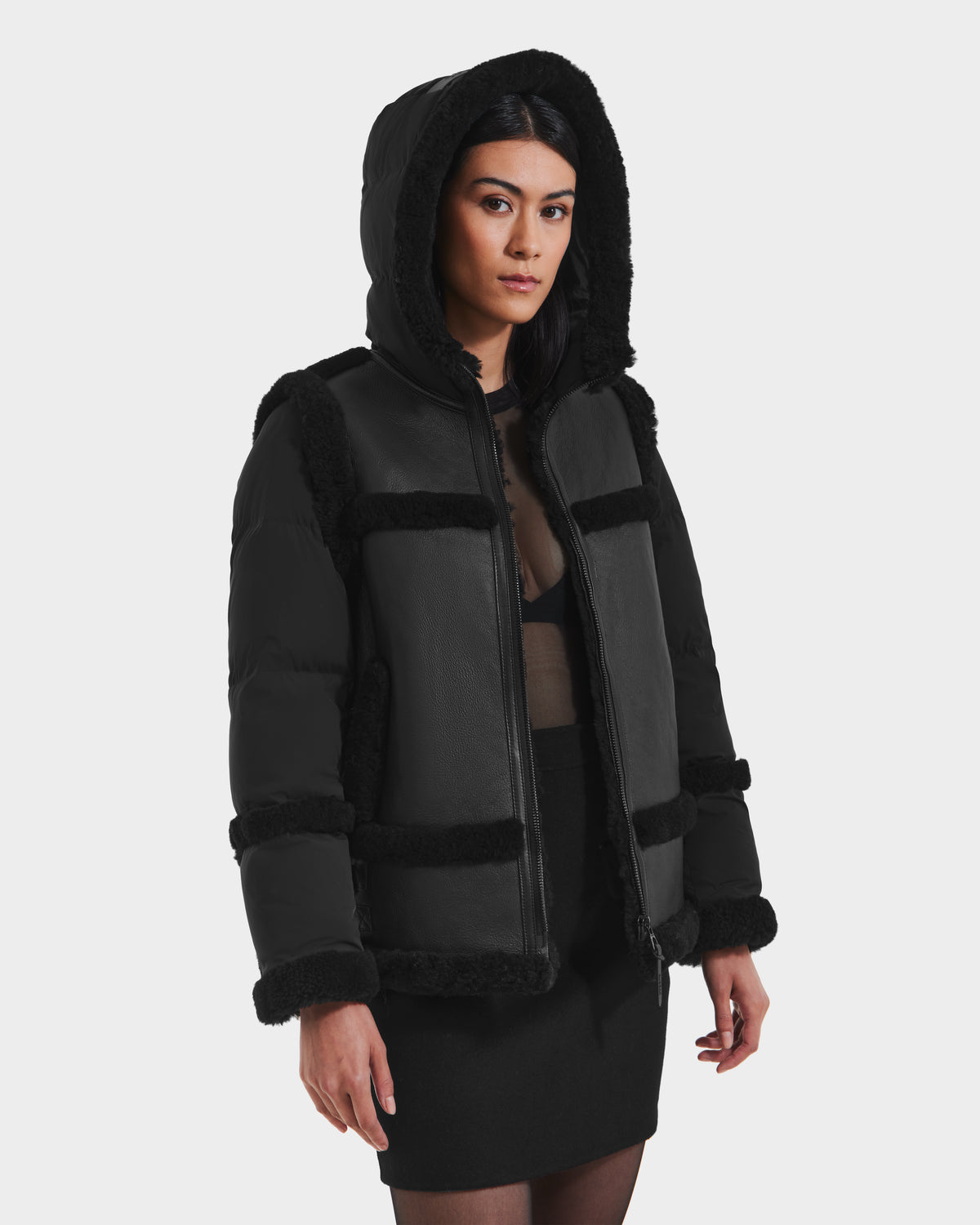 A woman wears the Rudsak PHEOBE - 8125510 Black women’s shearling puffer jacket over a sheer black top and skirt, standing against a white background and looking to the side with a neutral expression.