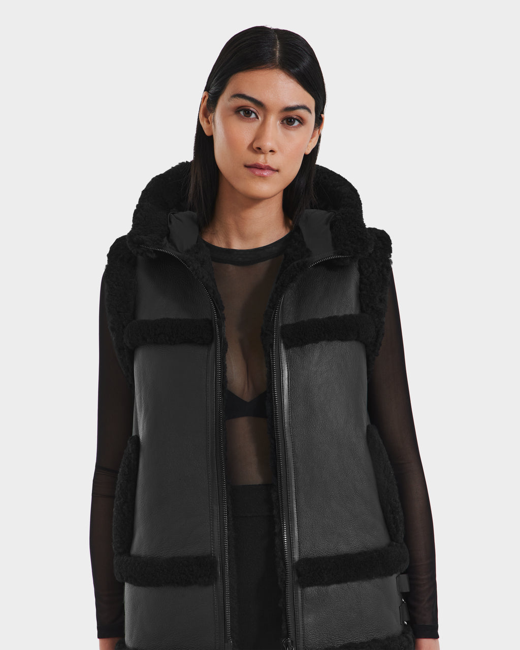 A woman with straight dark hair wears the Rudsak PHEOBE - 8125510 black shearling vest over a sheer black top, styled as a chic transitional jacket, standing against a plain light background and looking at the camera.