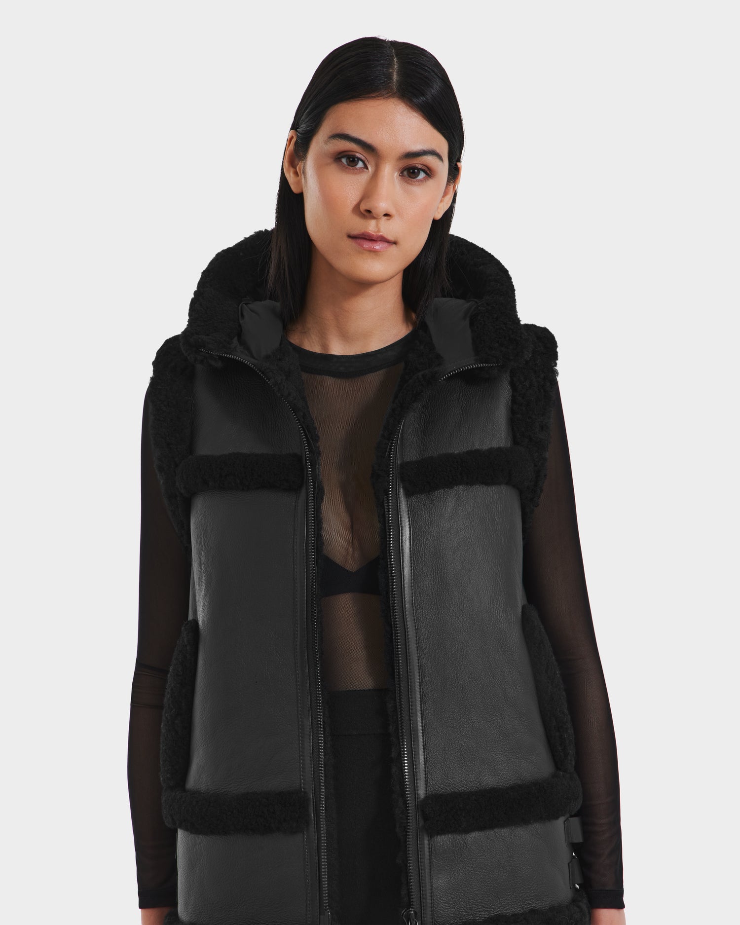 A woman with straight dark hair wears the Rudsak PHEOBE - 8125510 black shearling vest over a sheer black top, styled as a chic transitional jacket, standing against a plain light background and looking at the camera.