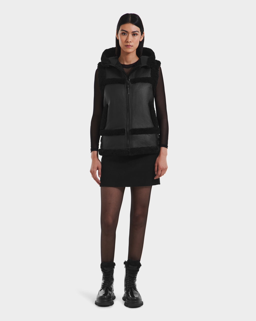 A woman faces forward against a white background, wearing the Rudsak PHEOBE - 8125510 Black transitional jacket over a black long-sleeve top, black skirt, sheer black tights, and black boots.
