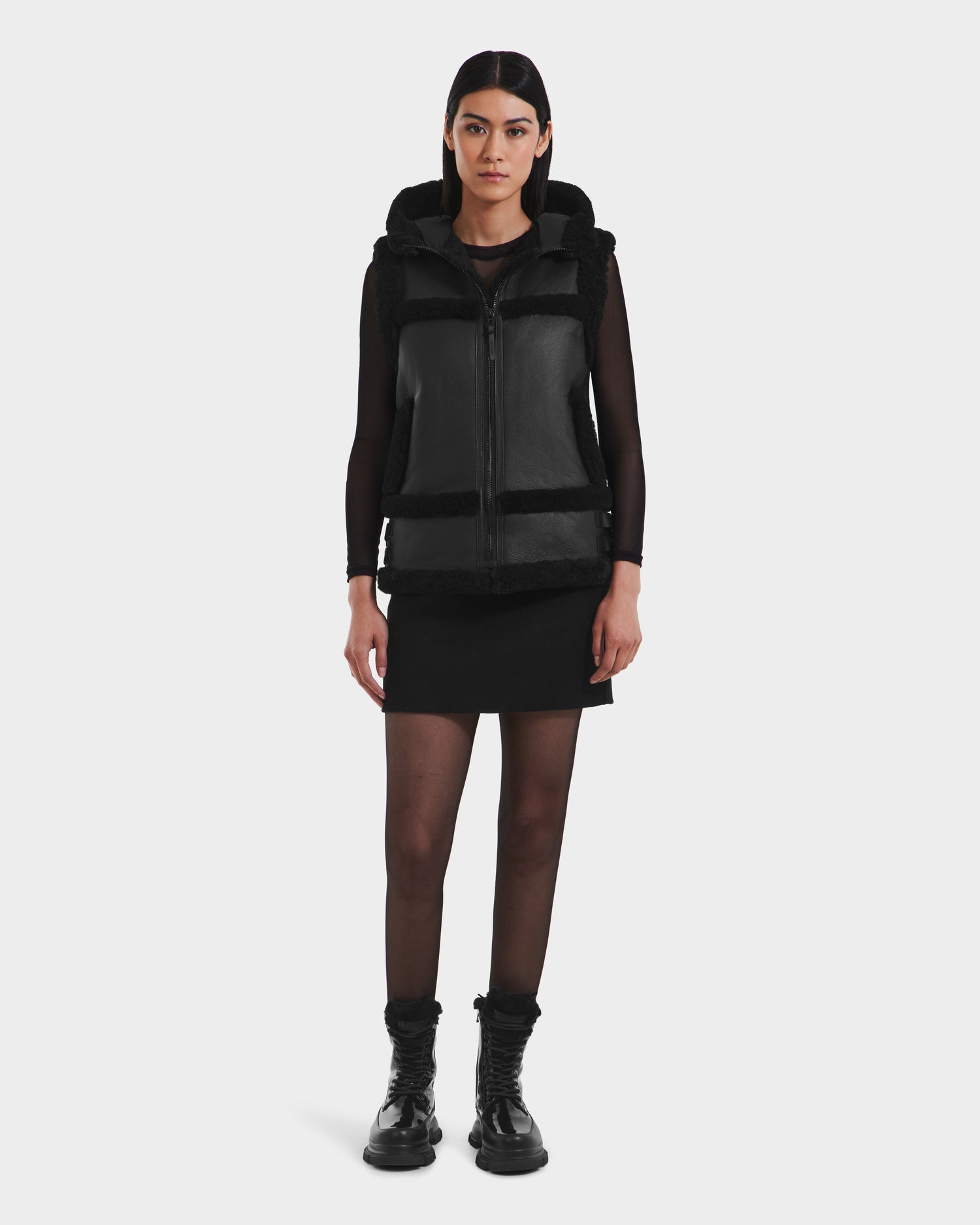A woman faces forward against a white background, wearing the Rudsak PHEOBE - 8125510 Black transitional jacket over a black long-sleeve top, black skirt, sheer black tights, and black boots.