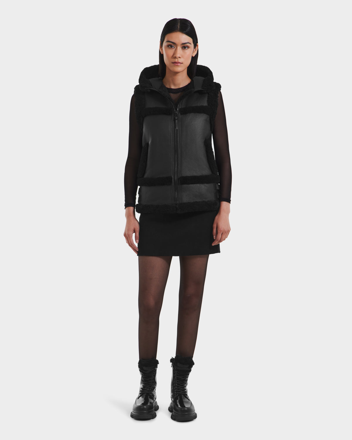 A woman faces forward against a white background, wearing the Rudsak PHEOBE - 8125510 Black transitional jacket over a black long-sleeve top, black skirt, sheer black tights, and black boots.