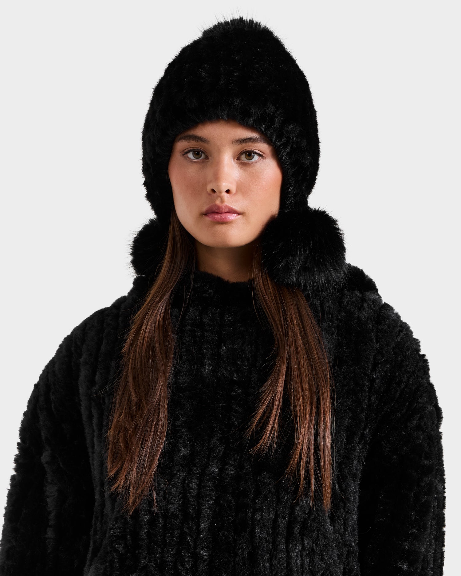 A young woman with long brown hair wears the Rudsak PIA - 8425547 Black women's faux fur hat with pom-poms and a matching coat, standing against a plain light background and looking directly at the camera with a neutral expression.