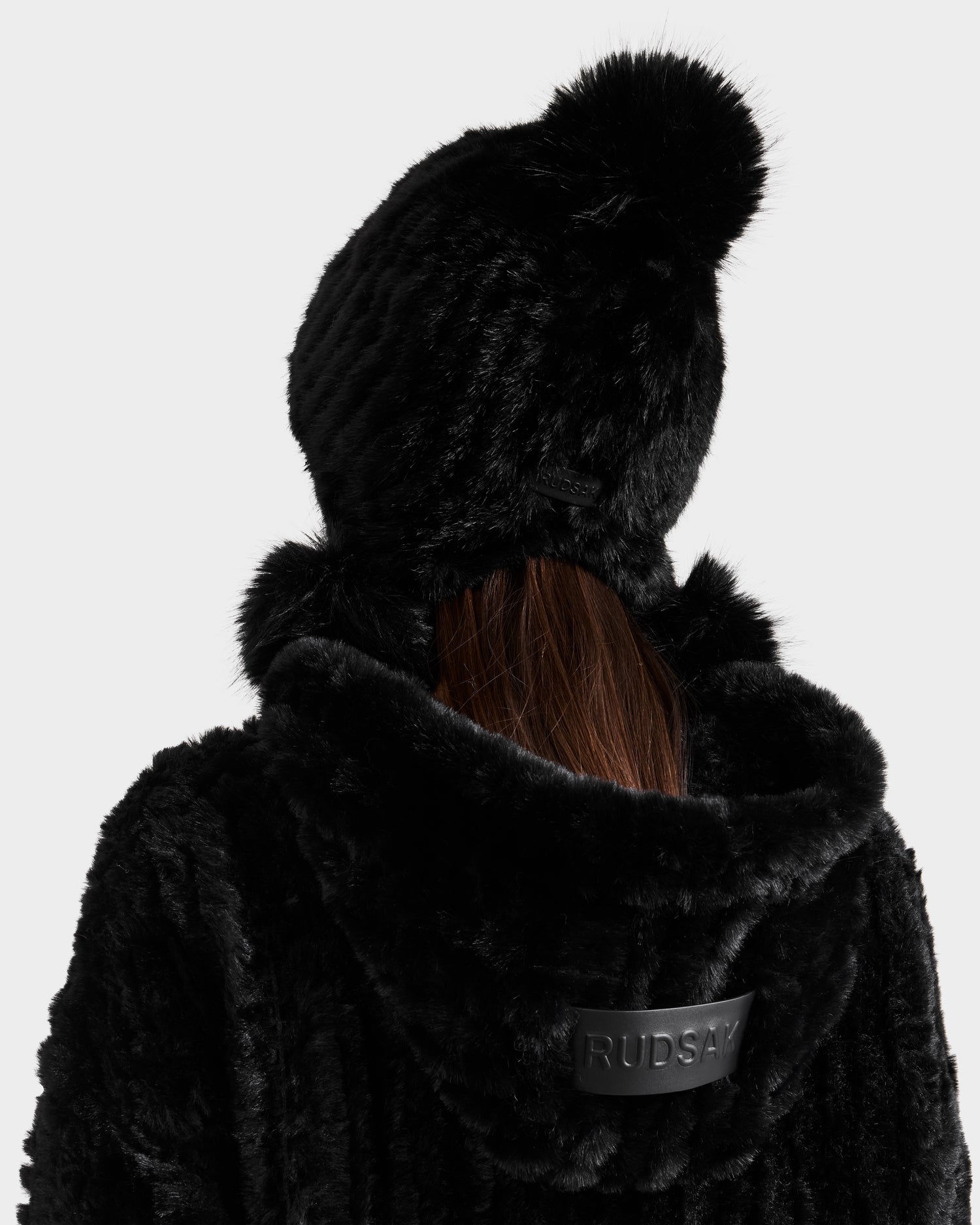 A person seen from behind wears the Rudsak PIA - 8425547 Black faux fur hooded coat and a matching black knitted pom-pom hat, making this stylish ensemble the perfect winter accessory.