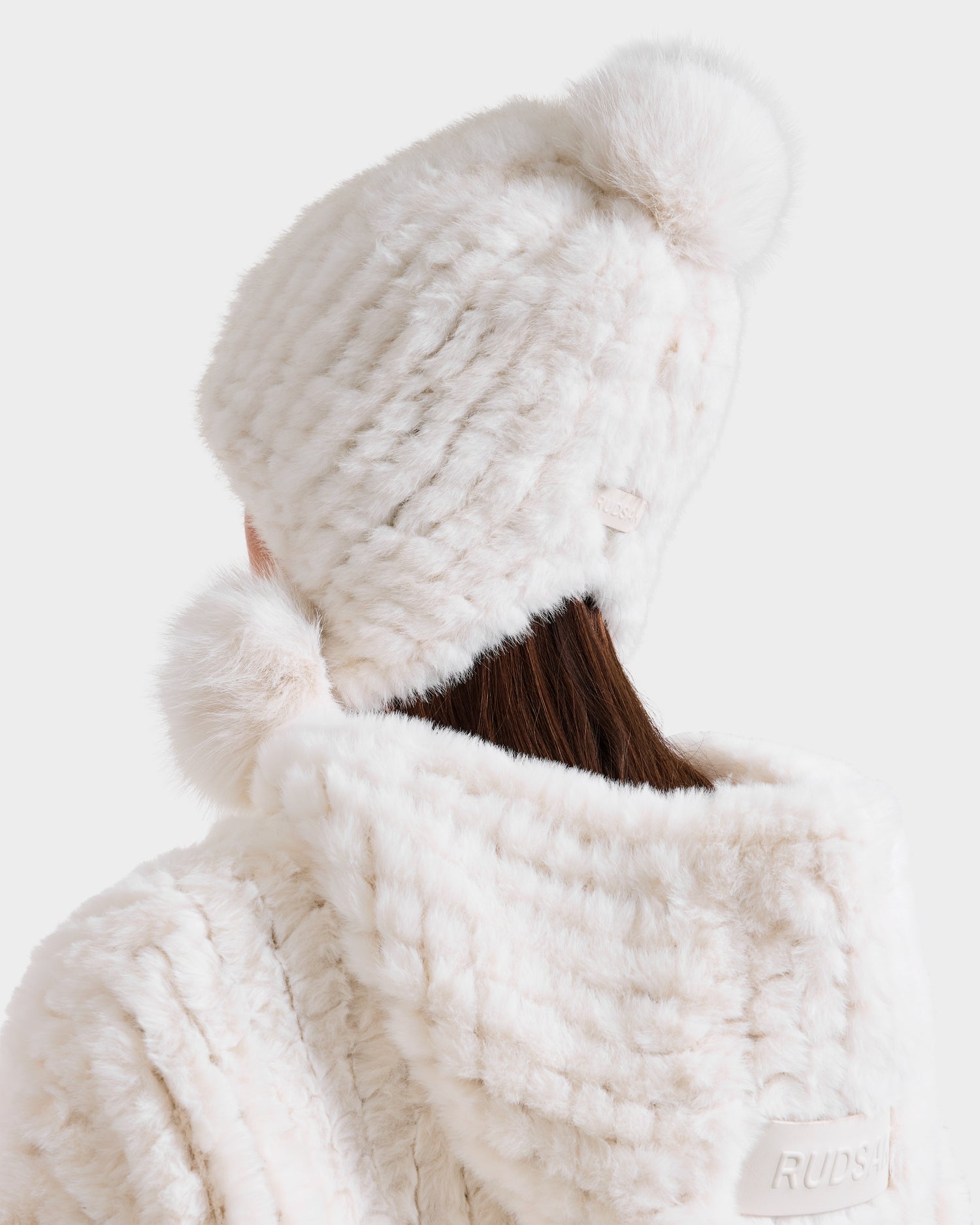 A person seen from behind wears the Rudsak PIA - 8425547 Iceland women’s faux fur hat with pom-poms and a matching faux fur coat with hood, creating a chic winter look against a plain white background.