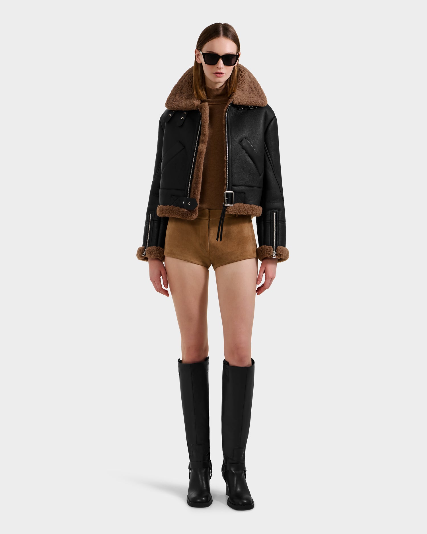 A woman wearing black sunglasses, a Rudsak POLINA - 8124811 Black women's shearling jacket, a brown turtleneck bodysuit, and knee-high black boots stands against a plain white background.