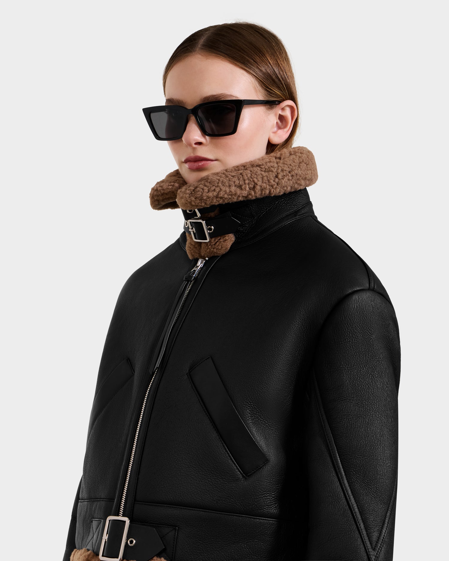A person poses against a plain light background wearing the Rudsak POLINA - 8124811 Black women's shearling jacket with a brown collar, large black sunglasses, and straight brown hair.