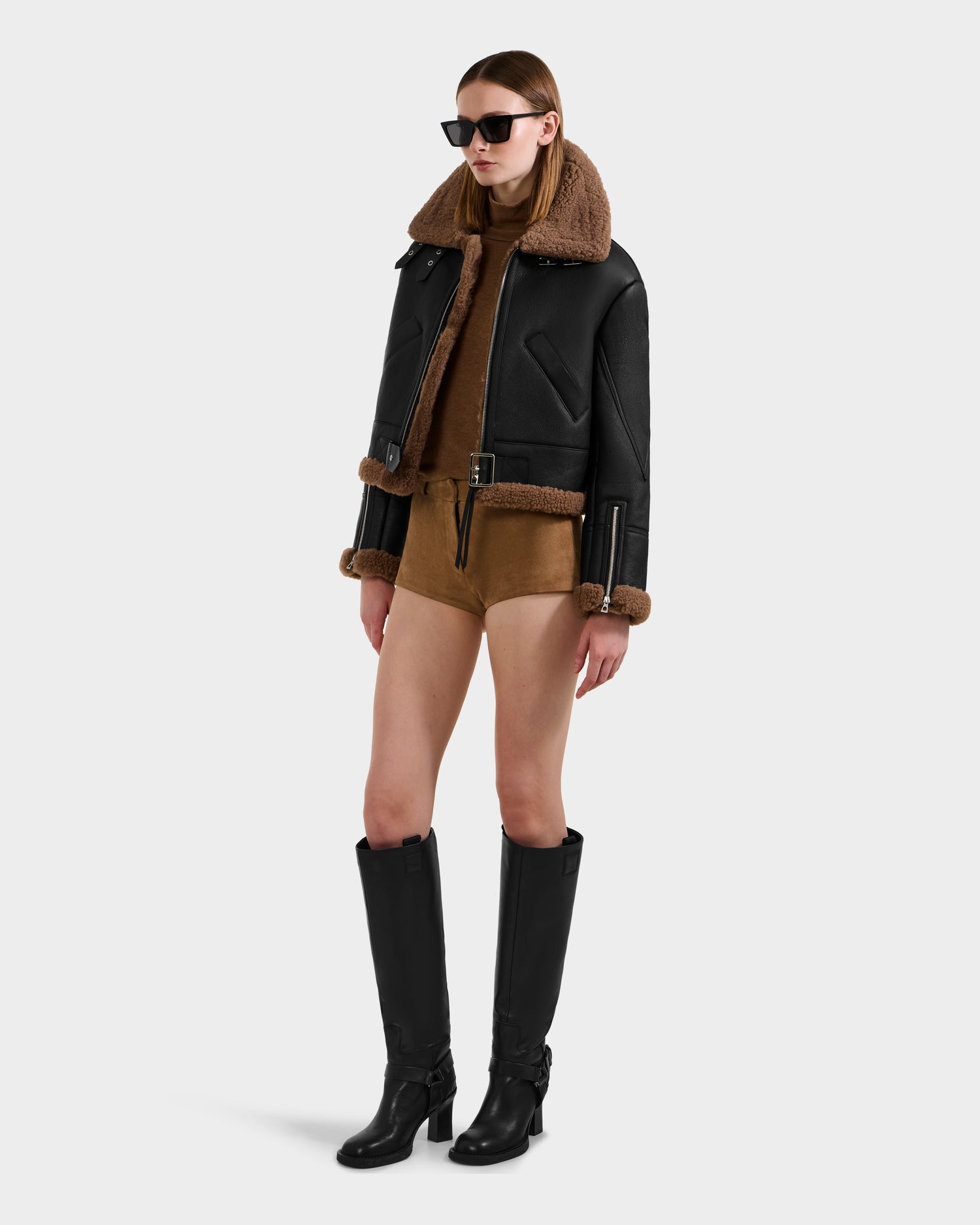A woman wearing black sunglasses, the Rudsak POLINA - 8124811 Black leather moto jacket with brown shearling trim, a brown outfit underneath, and tall black boots stands against a plain white background.