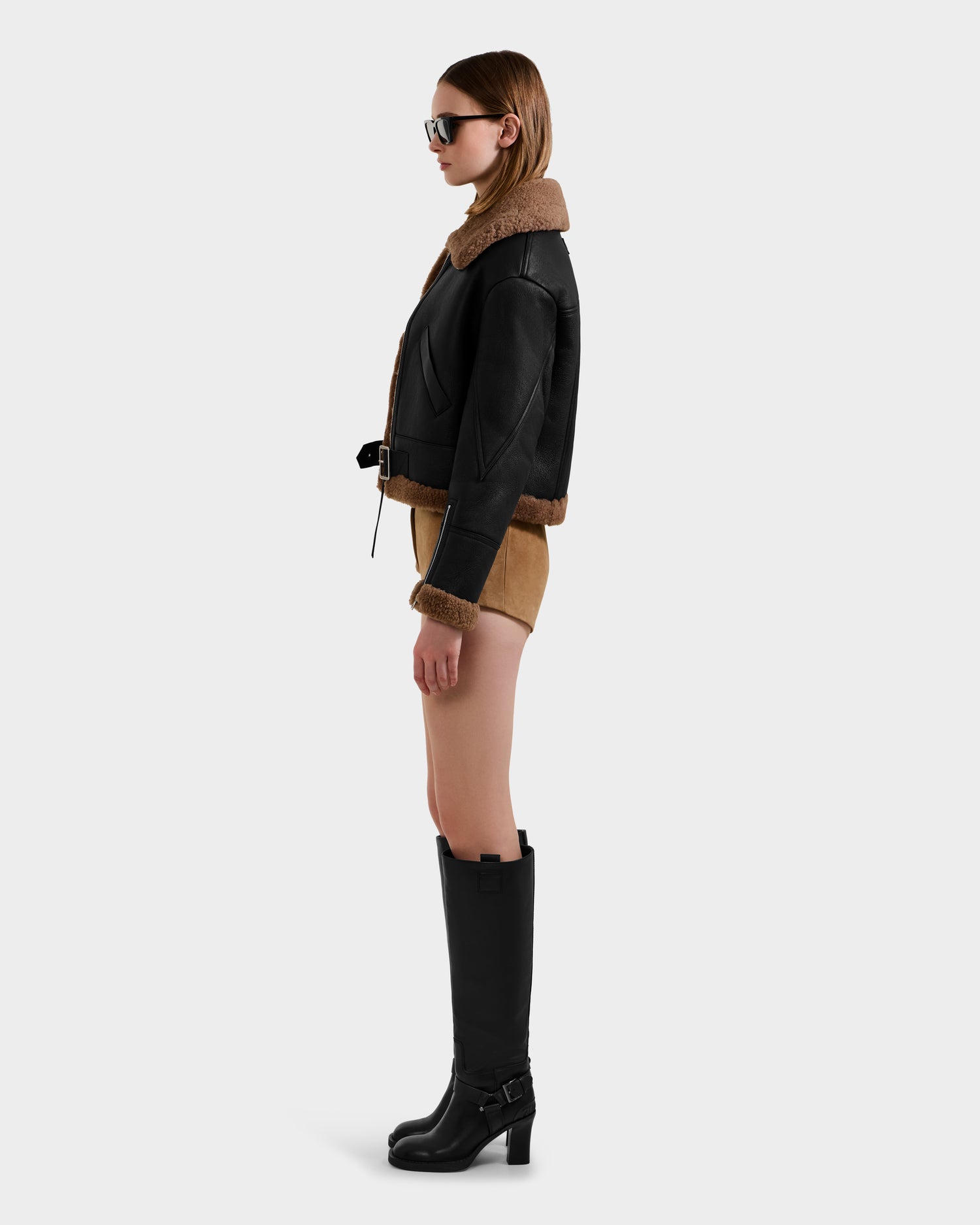 A woman in profile stands against a plain background, wearing sunglasses, tan shorts, knee-high black boots, and a chic Rudsak POLINA - 8124811 Black women's shearling jacket.