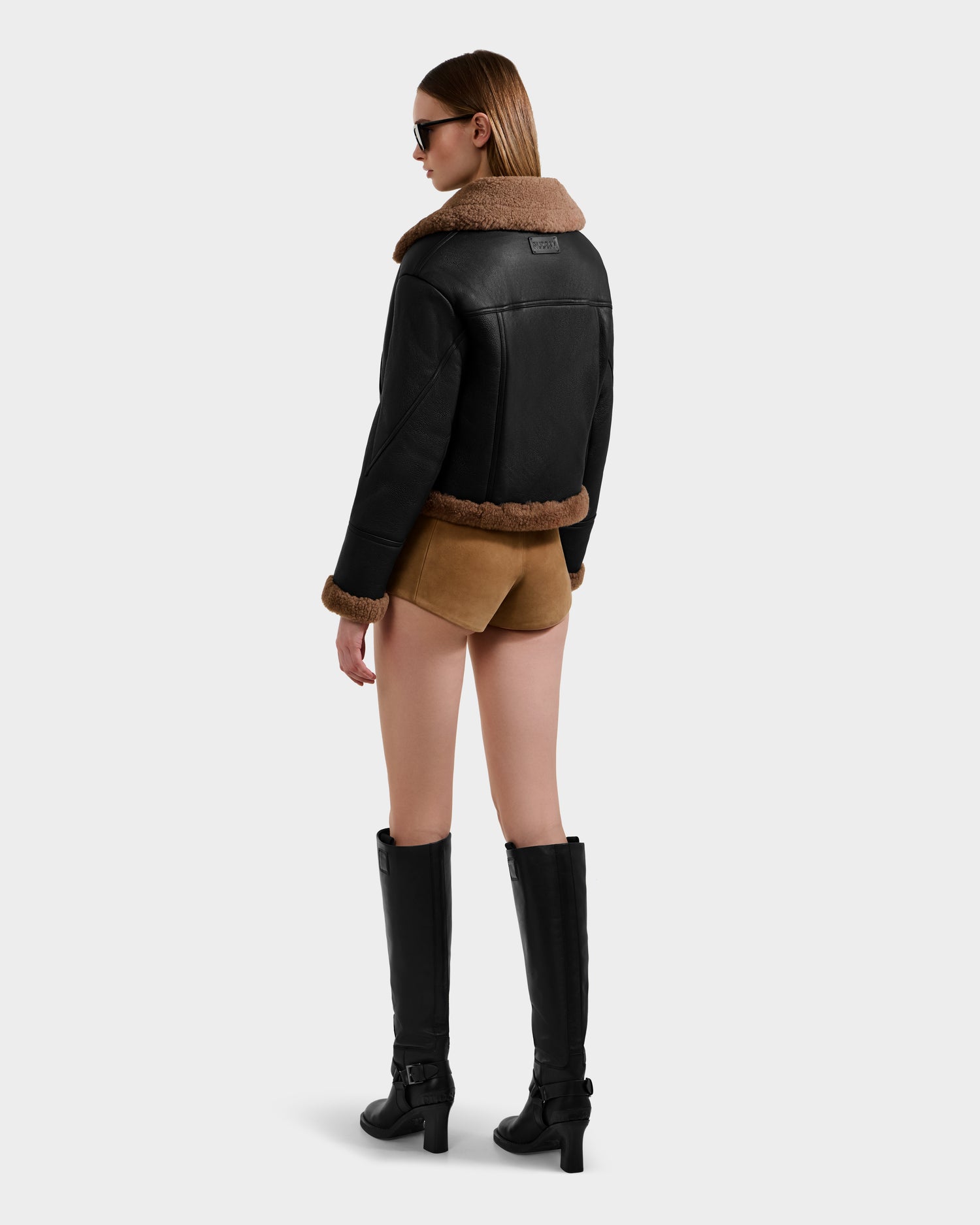 A woman faces away in sunglasses, wearing the Rudsak POLINA - 8124811 Black women's shearling jacket with brown trim, tan shorts, and knee-high black boots against a plain light background.