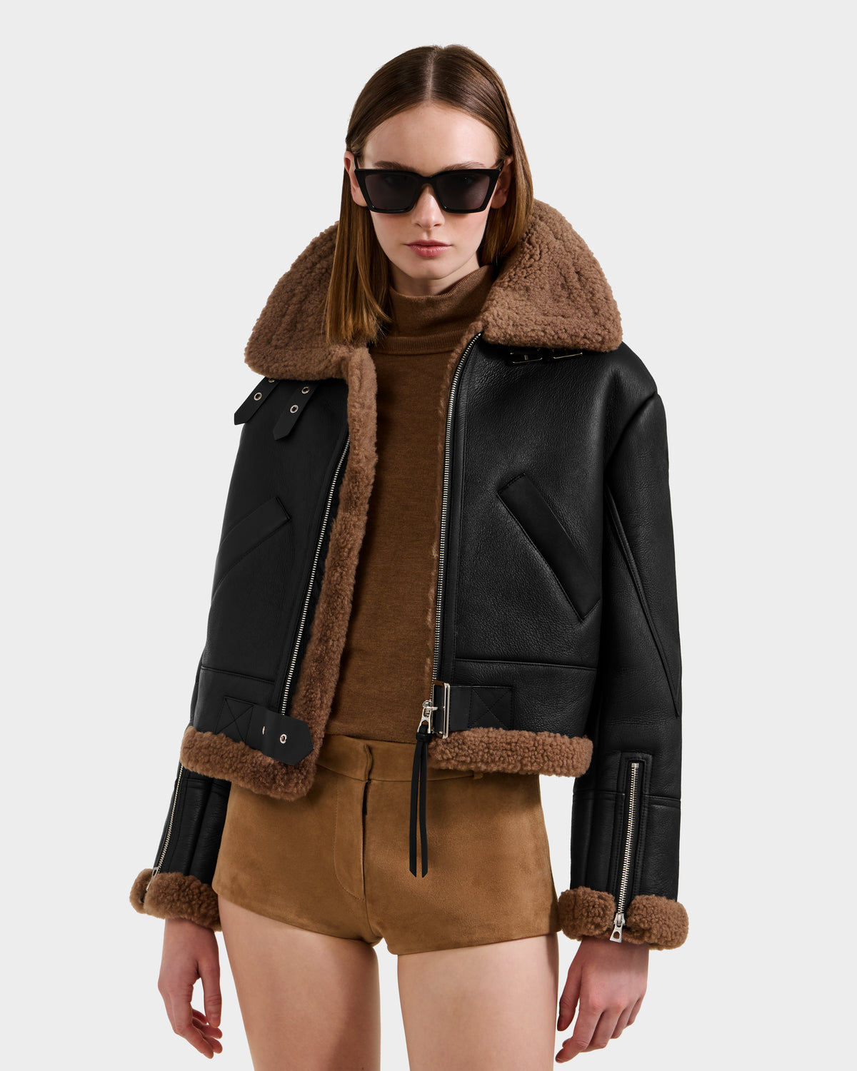 A woman in black sunglasses, a Rudsak POLINA - 8124811 Black leather moto jacket with brown shearling trim, a brown turtleneck sweater, and brown suede shorts stands against a plain light background.