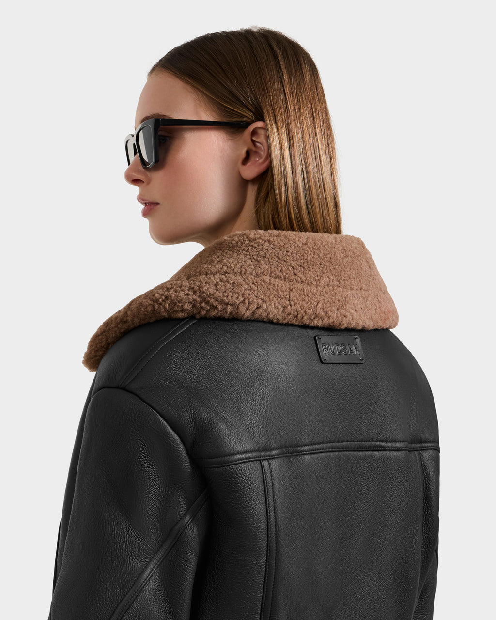 A woman in a Rudsak POLINA - 8124811 Black leather moto jacket with a tan shearling collar and sunglasses stands against a plain light background, facing away and glancing to the side.