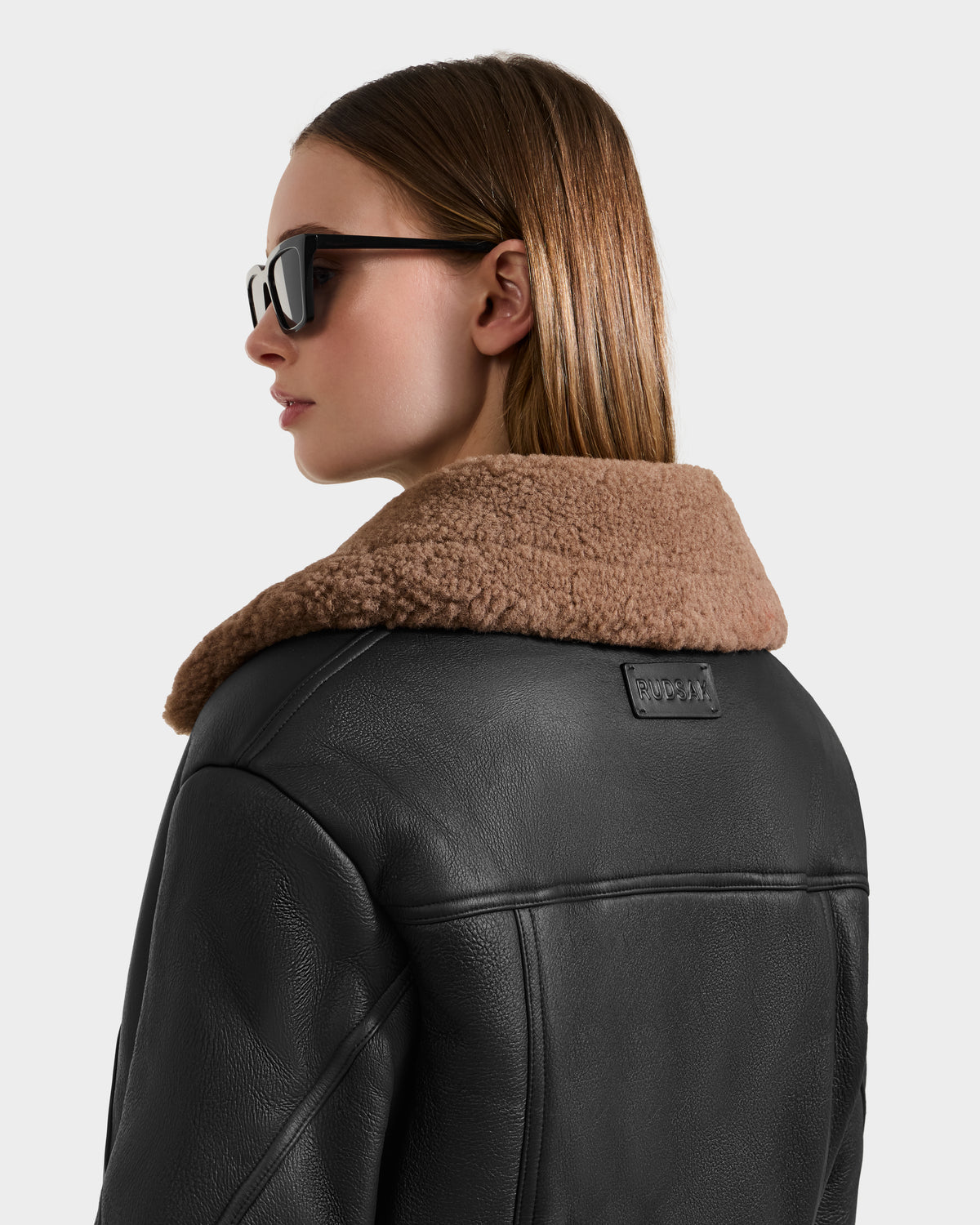 A woman in a Rudsak POLINA - 8124811 Black leather moto jacket with a tan shearling collar and sunglasses stands against a plain light background, facing away and glancing to the side.