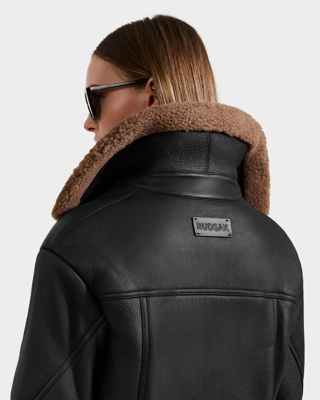 A person with straight, light brown hair and dark sunglasses wears a Rudsak POLINA - 8124811 Black leather moto jacket with a brown shearling collar, featuring the Rudsak label on the back, facing away from the camera.