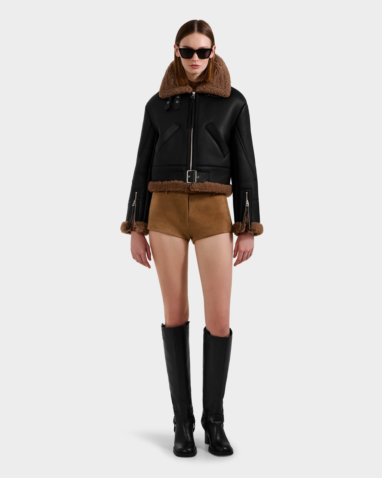 A person wearing the Rudsak POLINA - 8124811 Black women's shearling jacket, brown shorts, black knee-high boots, and dark sunglasses stands against a plain light gray background.