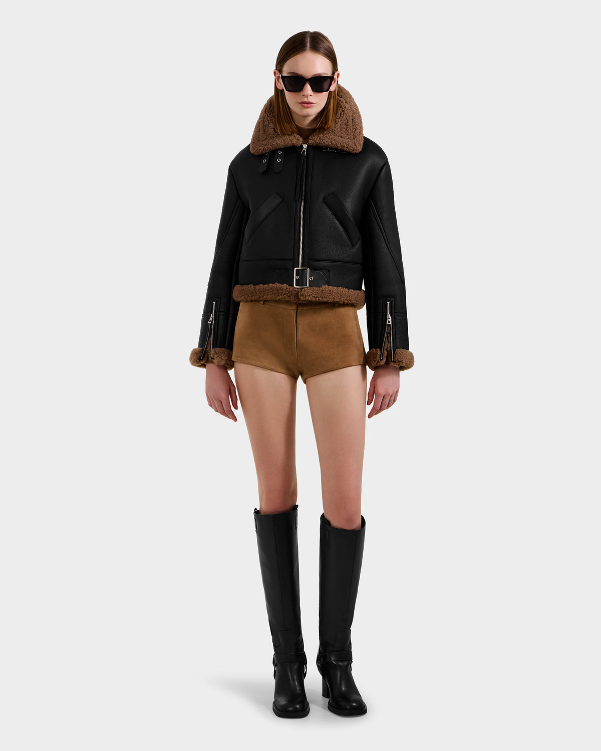 A person wearing the Rudsak POLINA - 8124811 Black women's shearling jacket, brown shorts, black knee-high boots, and dark sunglasses stands against a plain light gray background.
