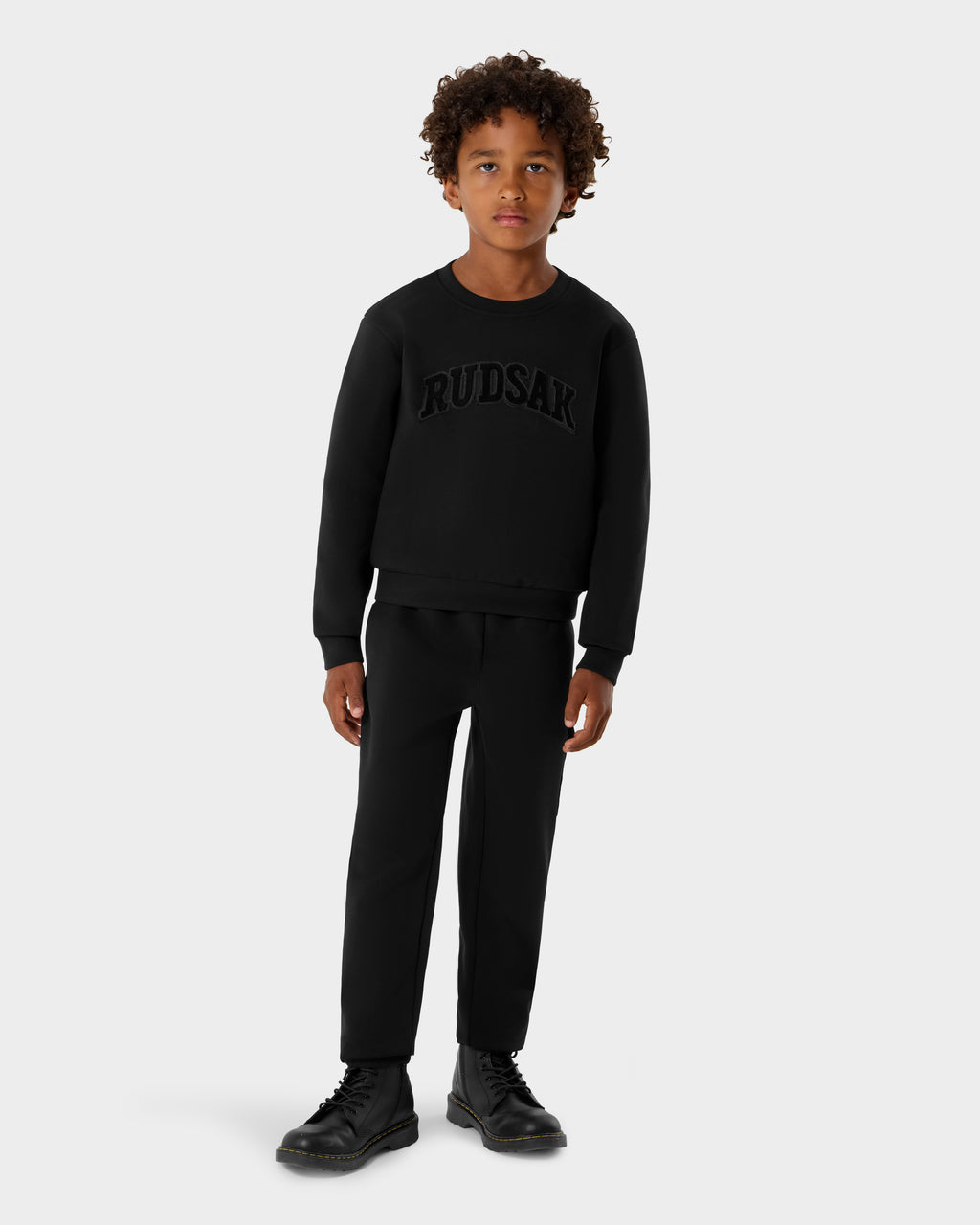 A young boy with curly hair stands against a plain light gray background, wearing a black Rudsak POPPY - 1525003 crewneck sweatshirt with matching kids’ black pants and black boots.
