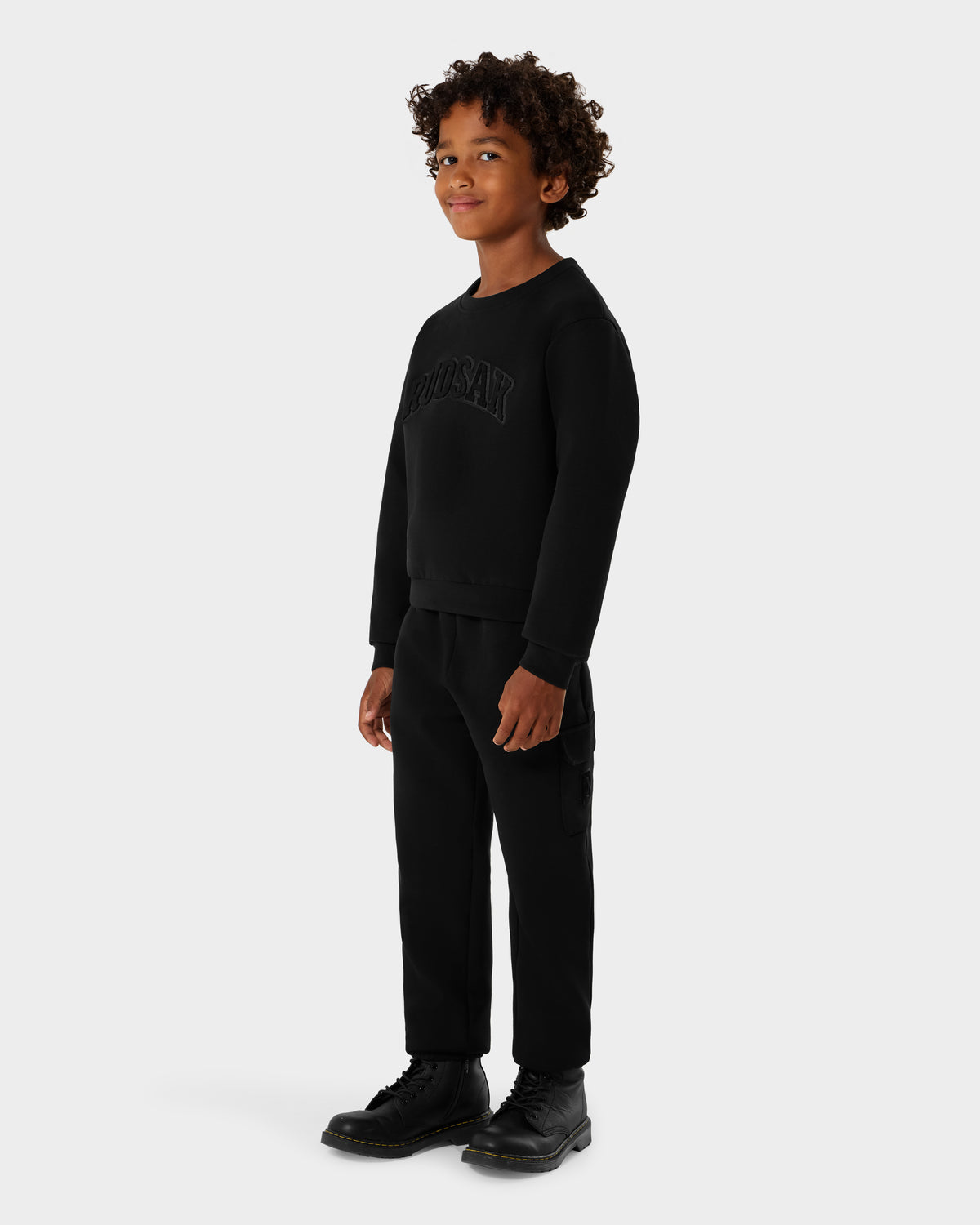 A young boy with curly hair stands slightly turned, smiling, in a black Rudsak POPPY - 1525003 sweatshirt with lettering, black pants, and black boots against a plain light gray background.