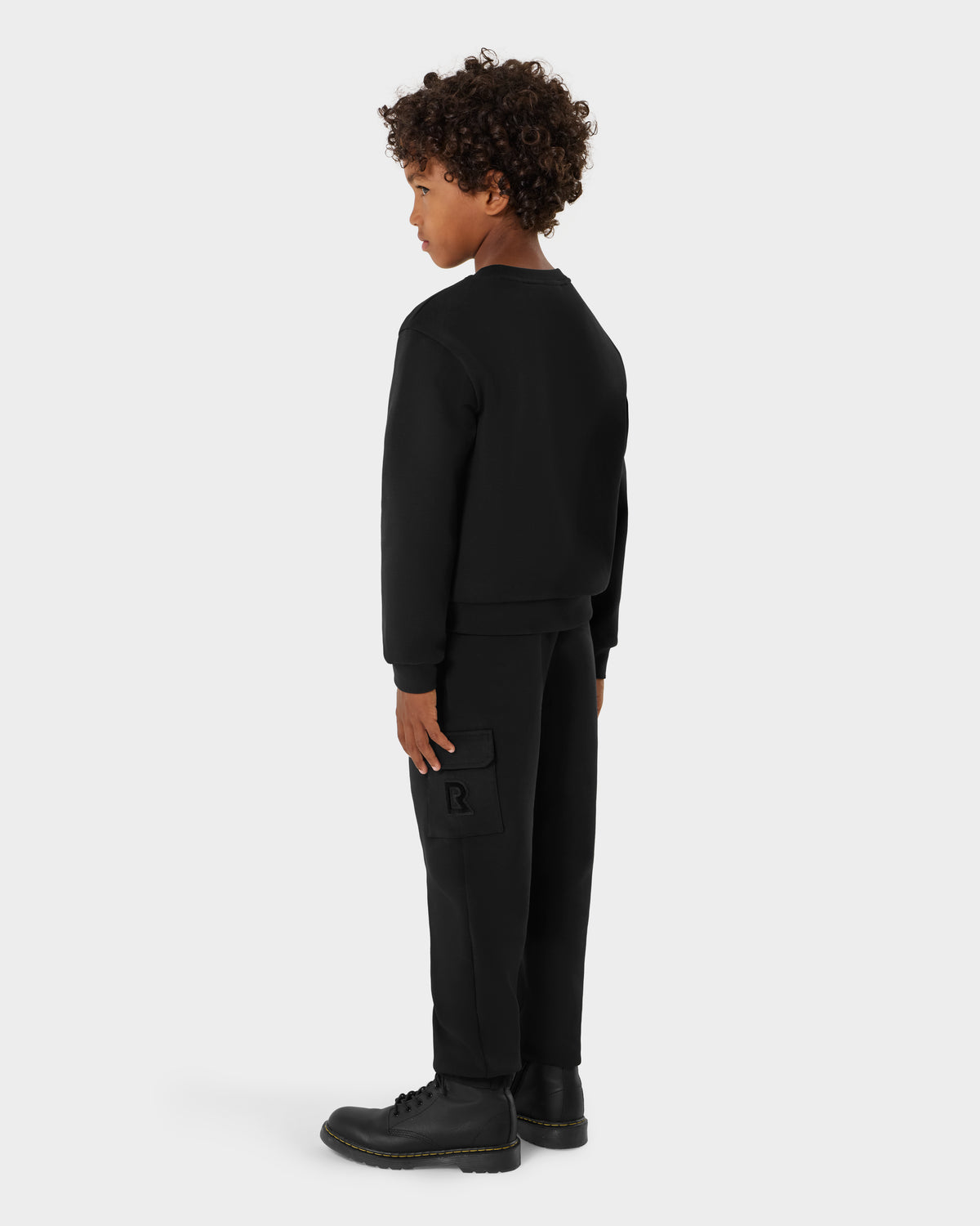 A young boy with curly hair wears the Rudsak POPPY - 1525003 Black crewneck sweatshirt, black cargo pants, and black boots, standing slightly turned against a plain light background. Stylish choice for fashion-forward kids.