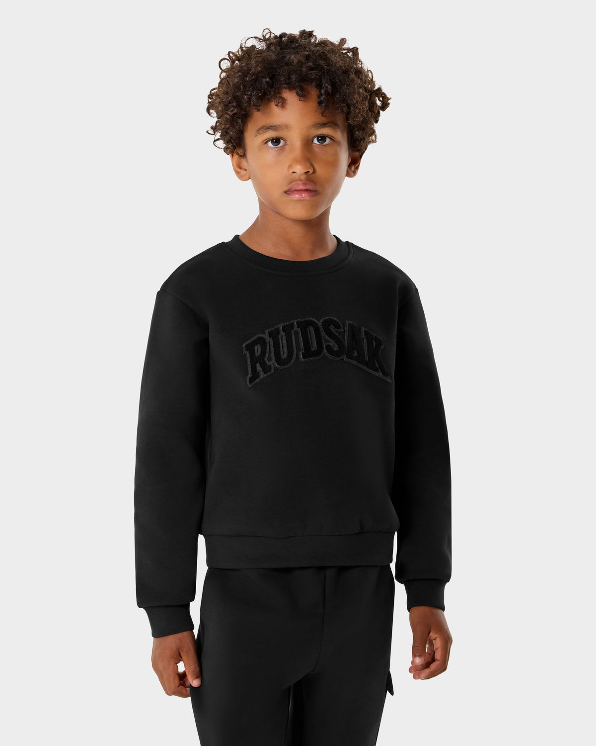 A young boy with curly hair wears the Rudsak POPPY - 1525003 Black crewneck and matching pants against a plain light gray background, offering kids the perfect blend of comfort and style.