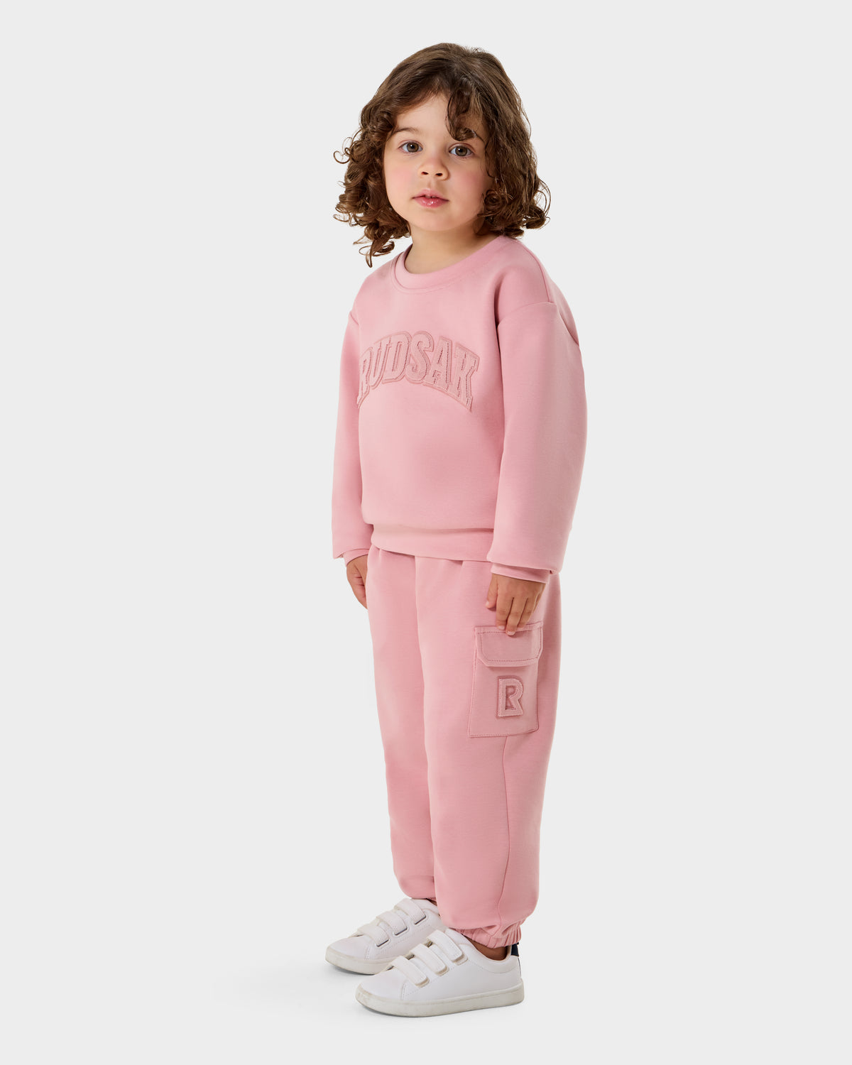 A young child with curly hair wears the Rudsak POPPY - 1525003 light pink premium cotton jersey crewneck sweatshirt and pants set, styled with white sneakers, posed against a plain white background.