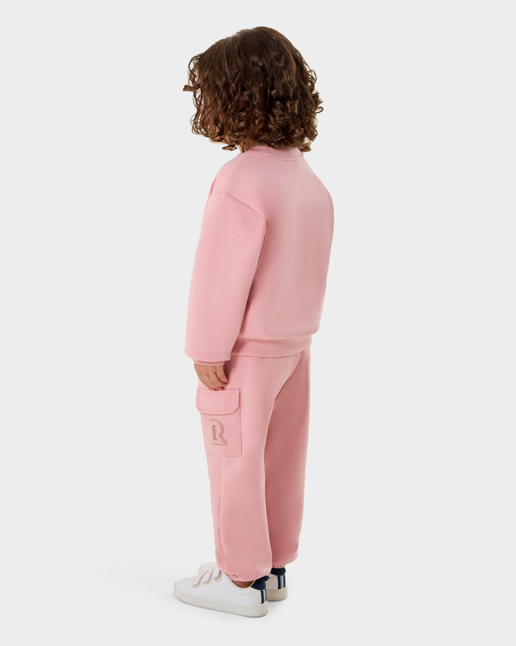 A child with curly brown hair, wearing the Rudsak POPPY - 1525003 light pink crewneck sweatshirt and matching pants made from premium cotton jersey, stands facing away in white sneakers with blue detail against a plain white background.