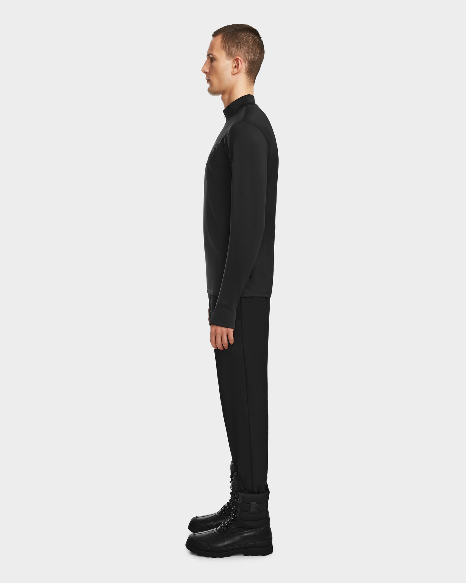 A man stands in profile against a white background, wearing the RALI - 6524522 Black men’s active top by Rudsak with black pants and lace-up boots—perfect for ski layering or winter sports.