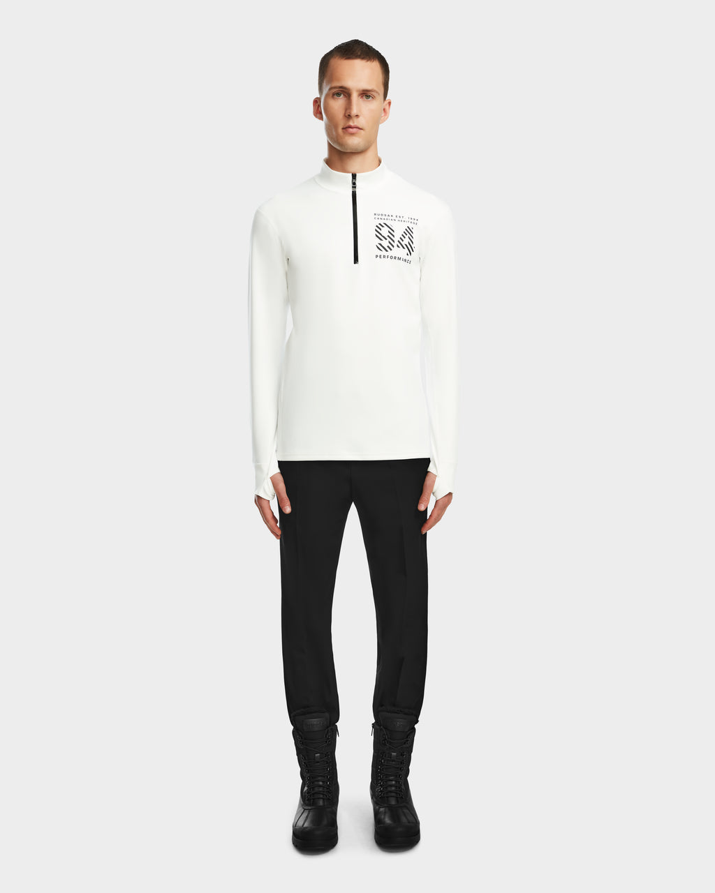 A man faces forward wearing the Rudsak RALI - 6524522 Optic White men’s active long-sleeve top with chest zipper and text graphic, paired with black pants and boots—perfect for ski layering—against a plain light background.