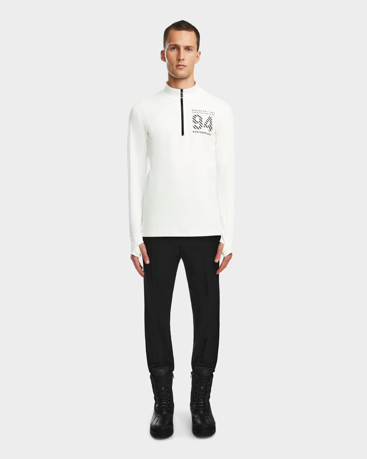 A man faces forward wearing the Rudsak RALI - 6524522 Optic White men’s active long-sleeve top with chest zipper and text graphic, paired with black pants and boots—perfect for ski layering—against a plain light background.