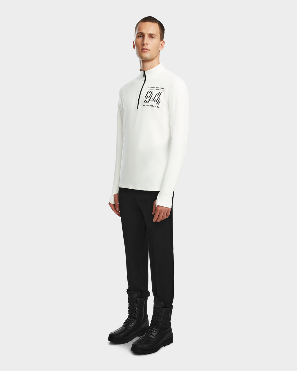 A man poses against a plain white background wearing the Rudsak RALI - 6524522 Optic White men’s long-sleeve zip-up featuring a black chest graphic, paired with black pants and lace-up boots—ideal for ski layering or winter sports.