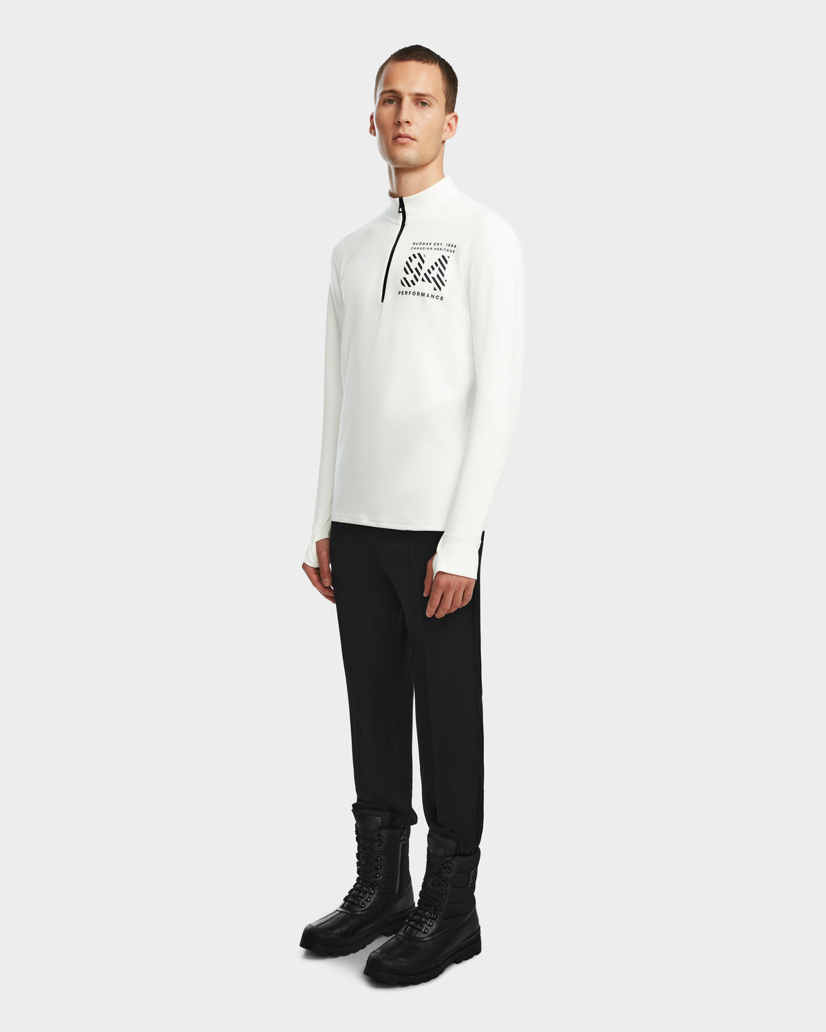 A man poses against a plain white background wearing the Rudsak RALI - 6524522 Optic White men’s long-sleeve zip-up featuring a black chest graphic, paired with black pants and lace-up boots—ideal for ski layering or winter sports.