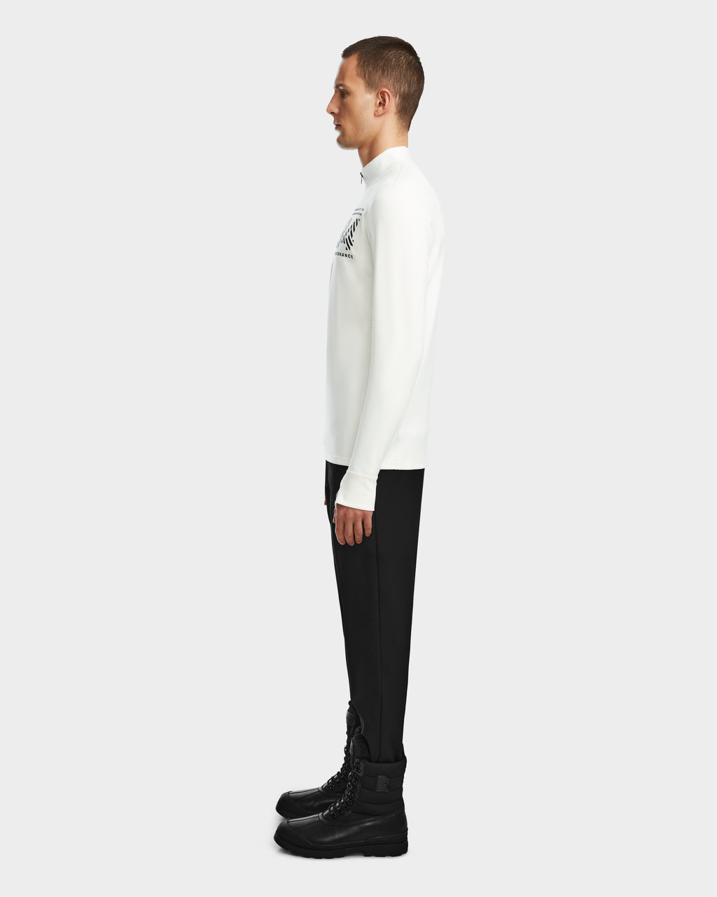 A man stands in profile facing left, wearing the RALI - 6524522 Optic White men’s active top by Rudsak, paired with black pants and lace-up boots—an ideal ski layering look—against a plain light background.