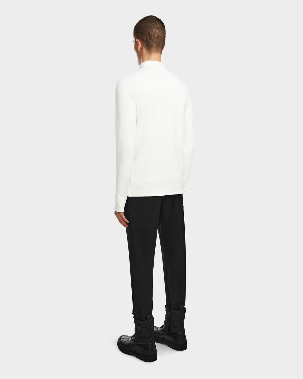 A person faces away, wearing the Rudsak RALI - 6524522 Optic White mens active top as a long-sleeve shirt with black pants and boots against a light gray background—ideal for winter sports or ski layering.