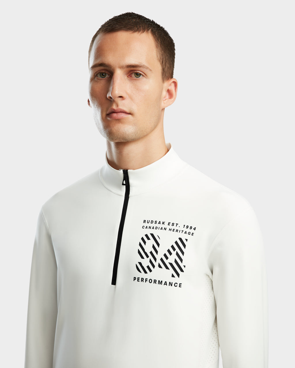 A man wears the RALI - 6524522 Optic White men’s active top by Rudsak, featuring black “RUDSAK EST. 1994 CANADIAN HERITAGE SW PERFORMANCE” text on the chest—ideal as a winter sports base layer against a light background.