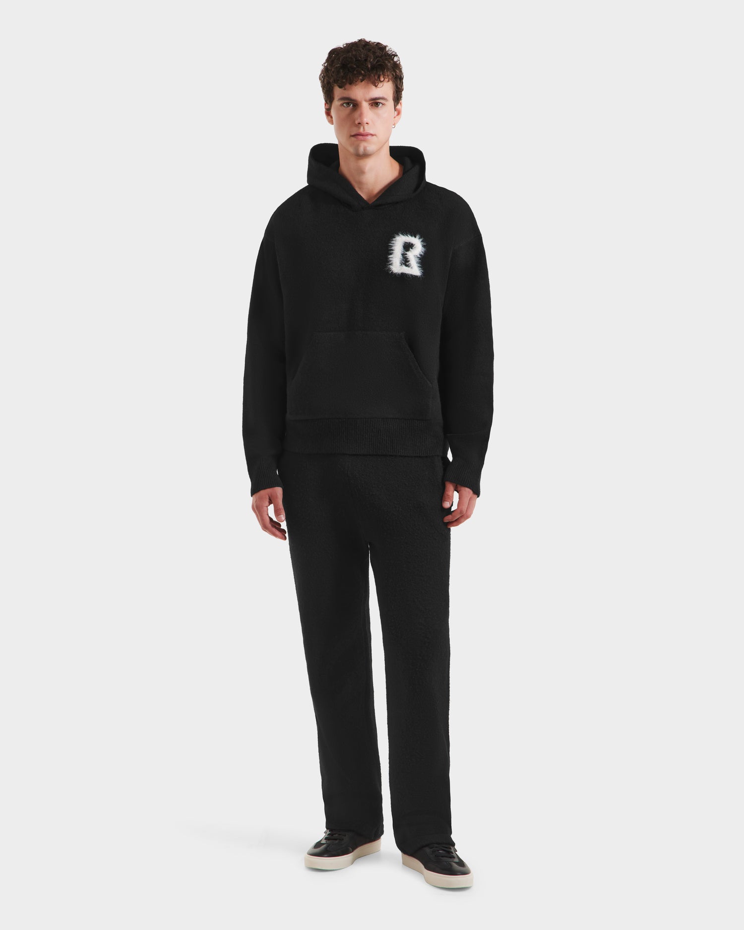 A young man faces forward in stylish streetwear—a Rudsak RAPHAEL - 6525643 Black men’s hoodie with a fuzzy white “R” on the chest, matching black pants, and black and white sneakers—set against a plain white background.